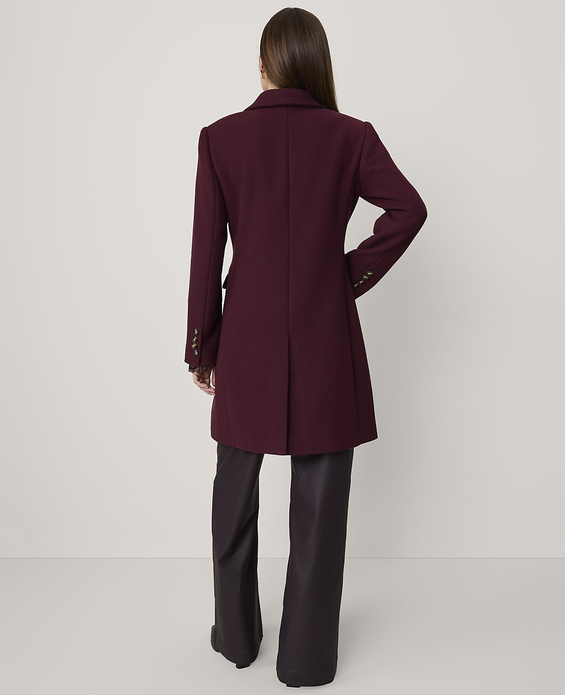 A woman stands with her back to the camera wearing a burgundy coat and dark wide-leg trousers against a plain light grey background.