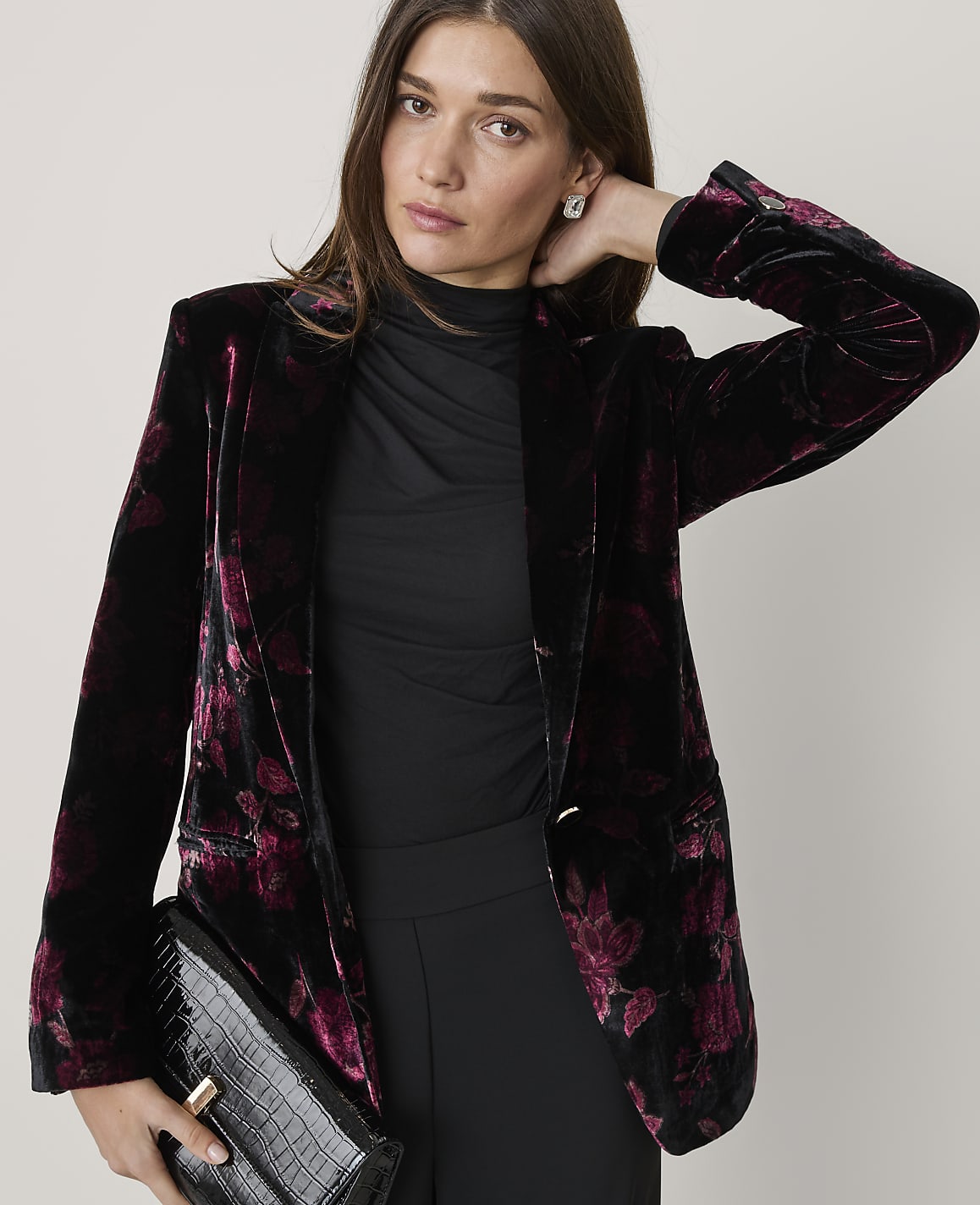 A woman wearing a floral velvet blazer and black clothing poses against a plain, light-colored background.
