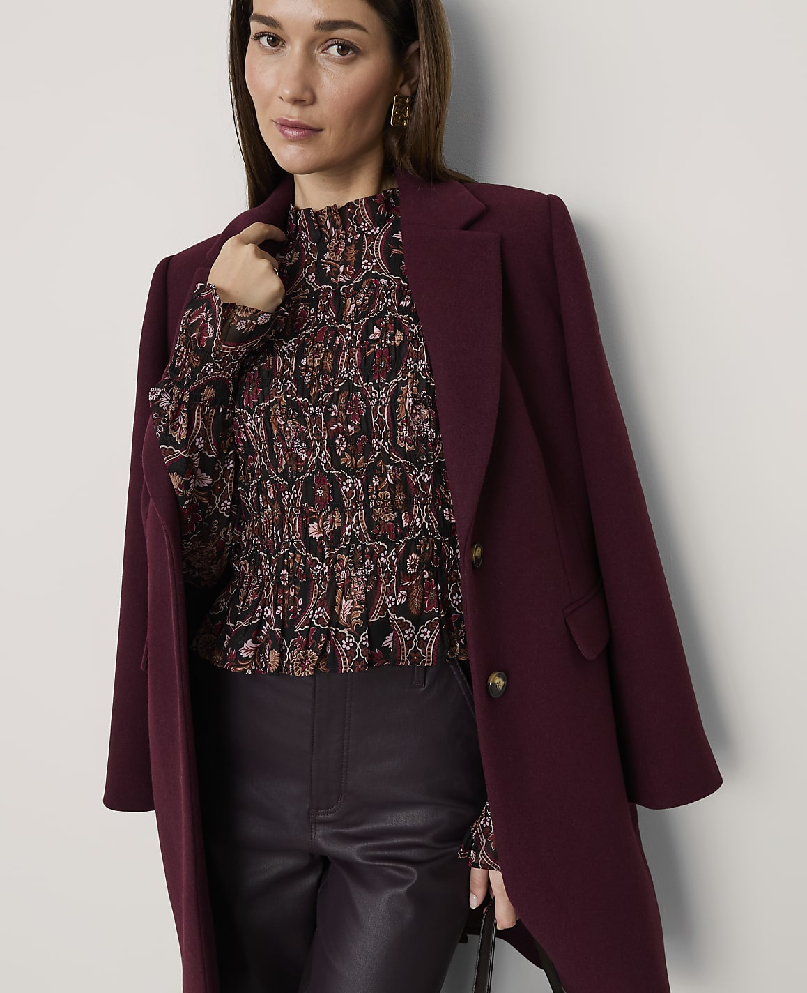 A woman wearing a patterned blouse, dark leather trousers, and a burgundy coat stands against a plain white background.