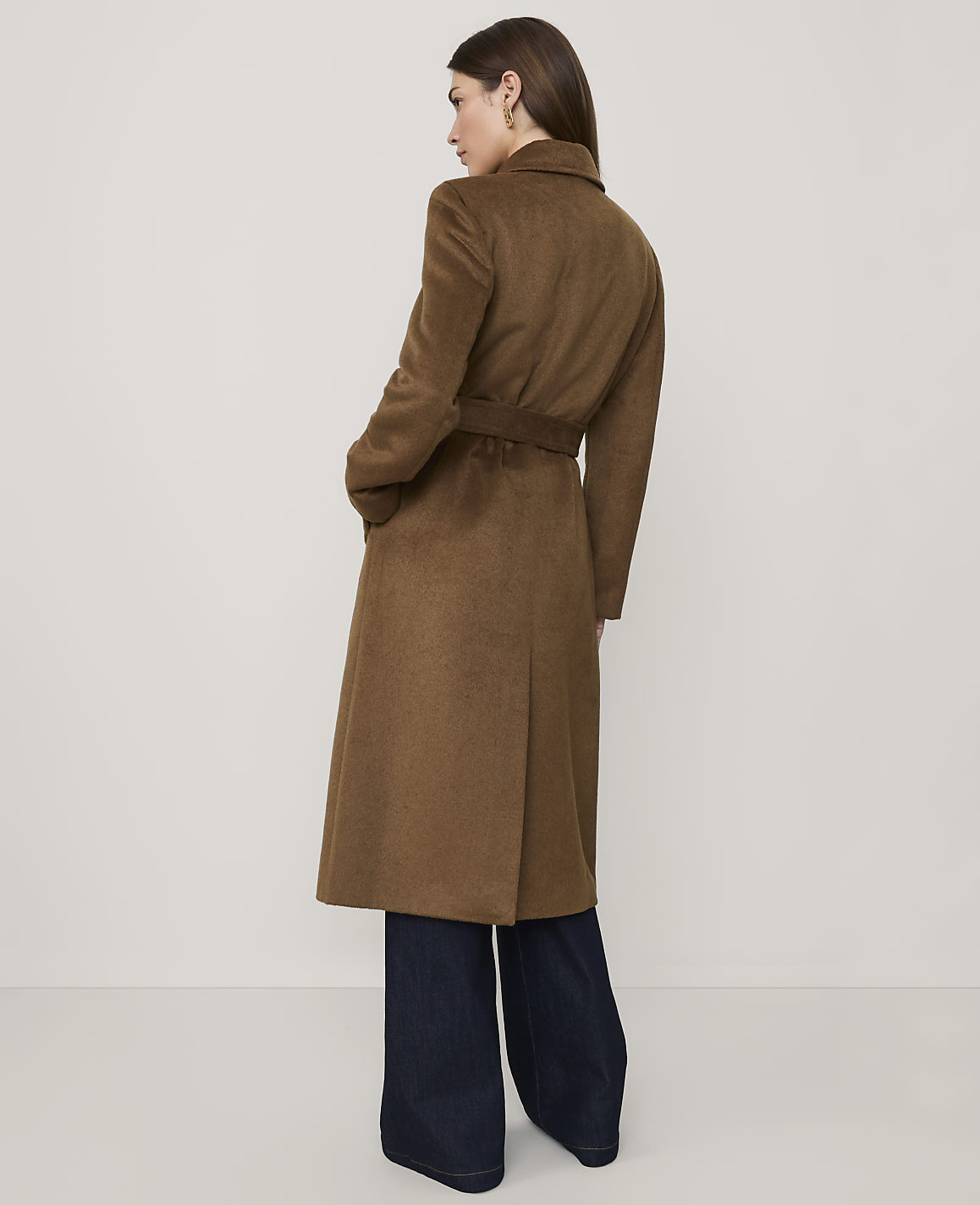 A woman wearing a long brown belted coat and wide-leg dark denim jeans stands against a plain, light-colored background.