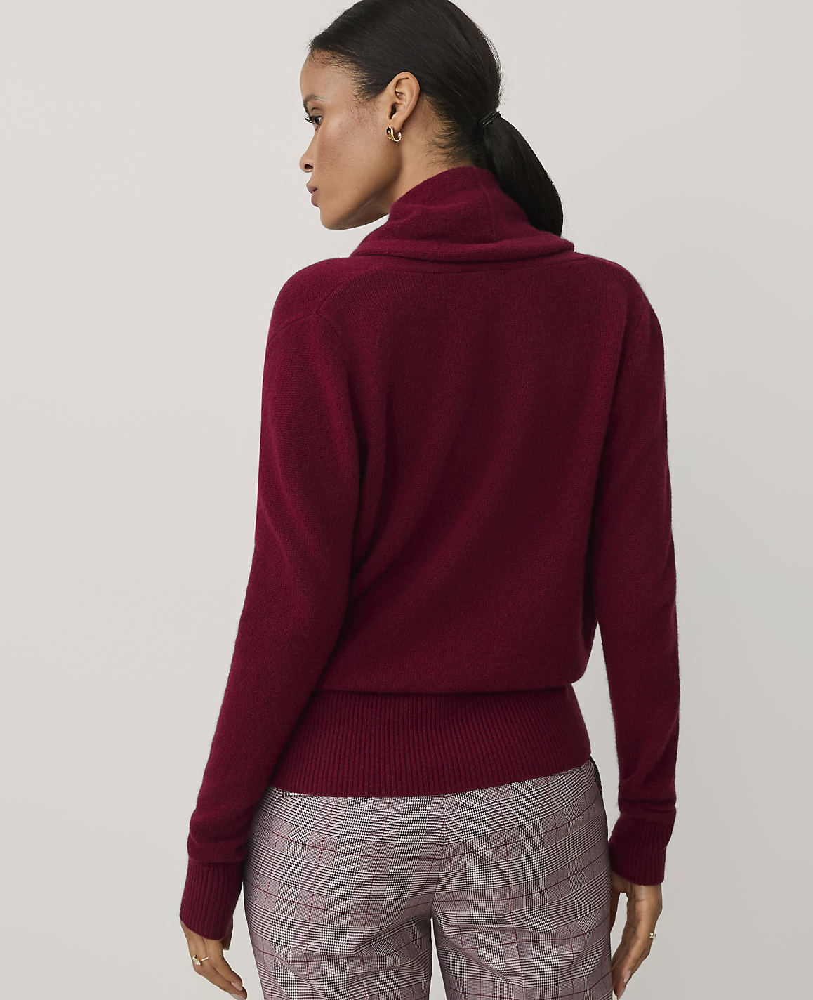 A woman wearing a burgundy turtleneck sweater and plaid trousers stands against a plain light gray background.