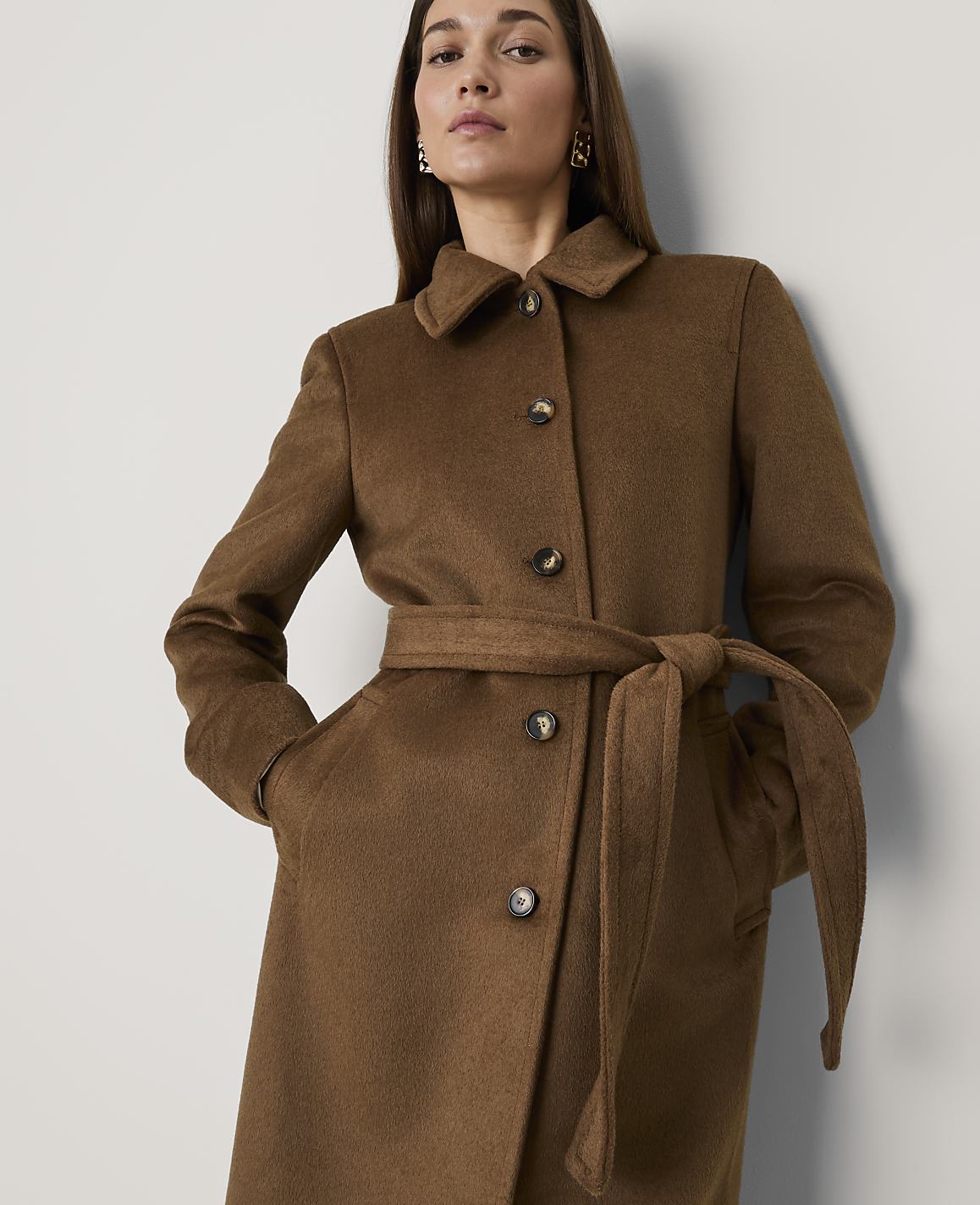 A woman wearing a brown belted coat stands against a plain white wall.