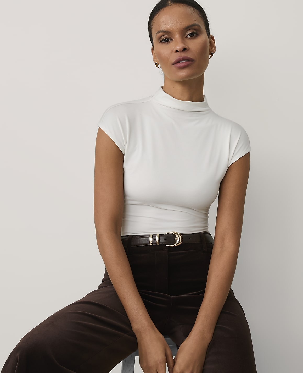 A woman wearing a white high-neck top and brown trousers sits against a plain, light-colored background.