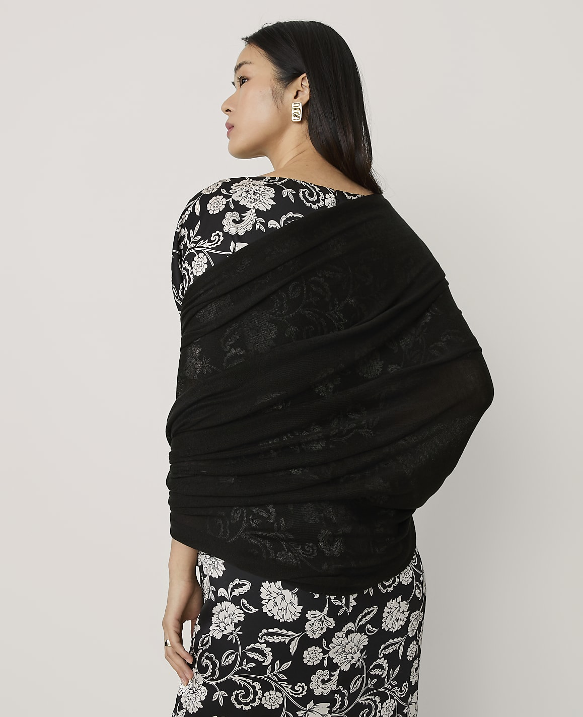 A woman wearing a black and white floral dress and a black shawl stands against a plain light gray background.