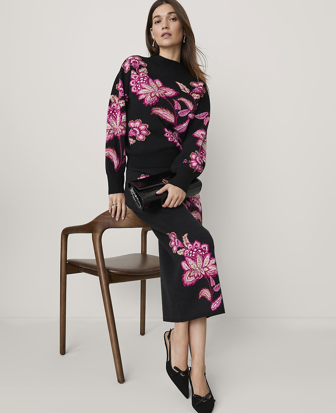 A woman wearing a black knit set with pink floral embroidery stands beside a wooden chair against a plain light grey background.