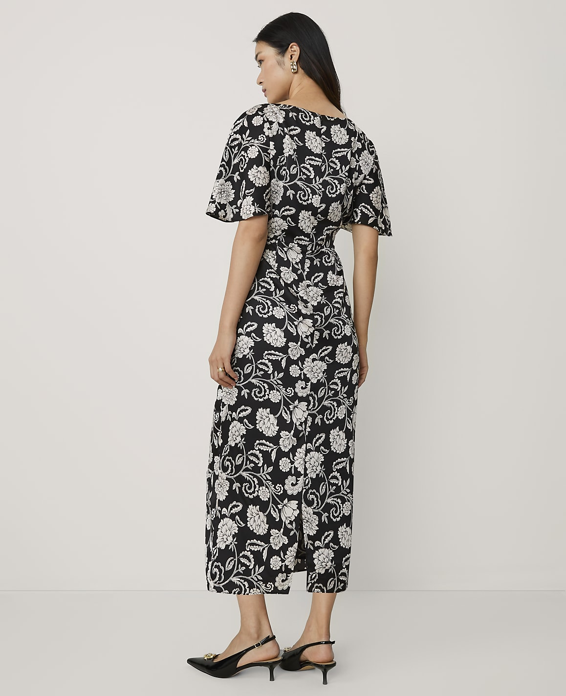 A woman wearing a black and white floral midi dress and black heels stands against a plain white background.