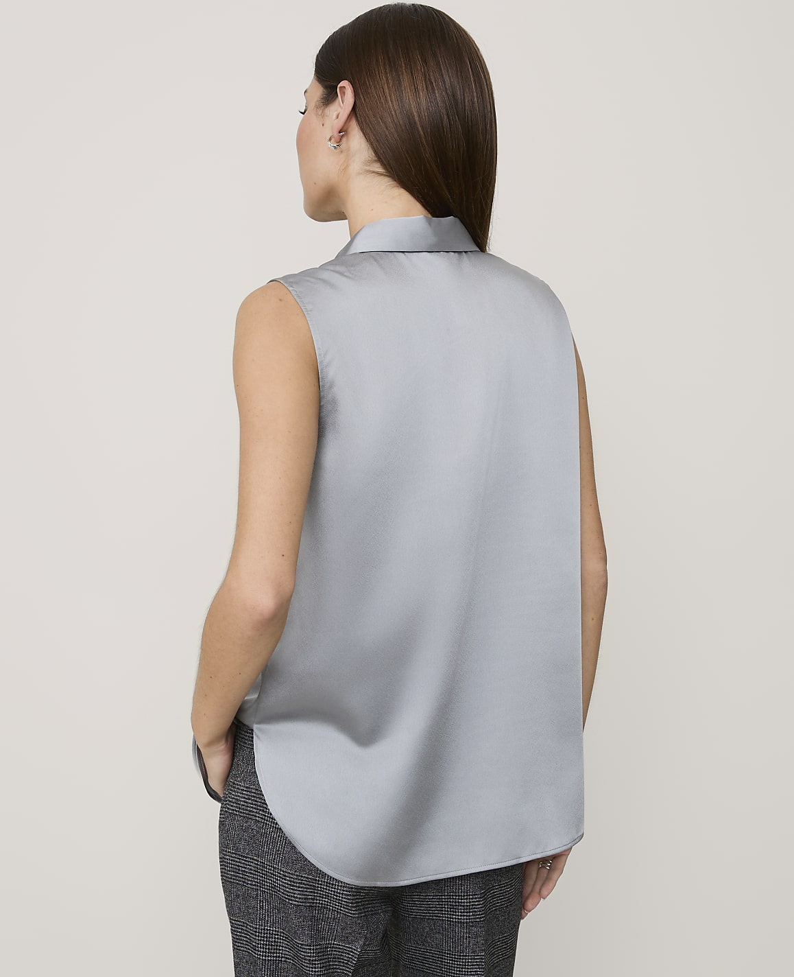 A woman wearing a sleeveless grey satin blouse and dark patterned trousers stands against a plain light grey background.