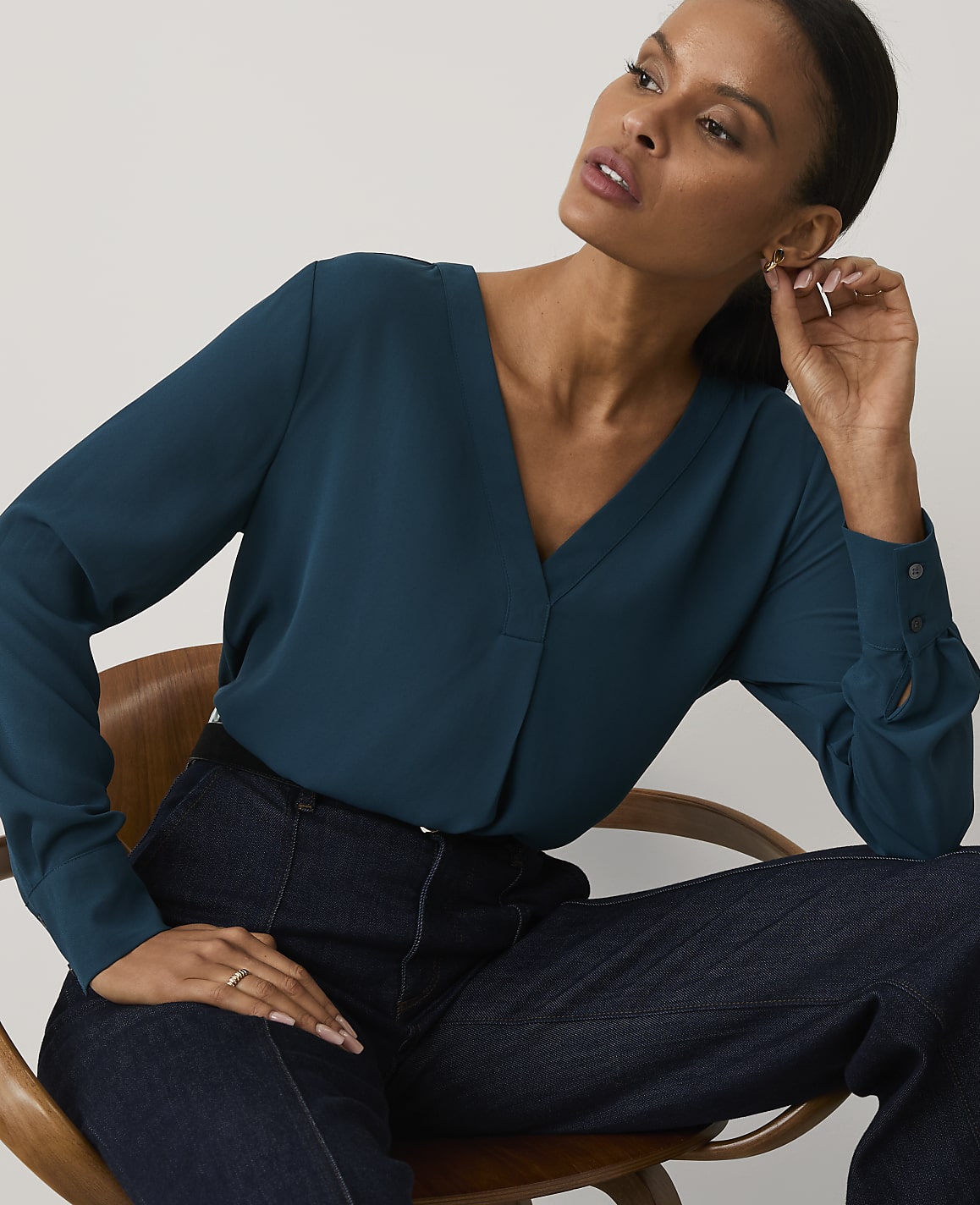 A woman wearing a teal V-neck blouse and dark blue jeans sits in a wooden chair against a plain, light-colored background.