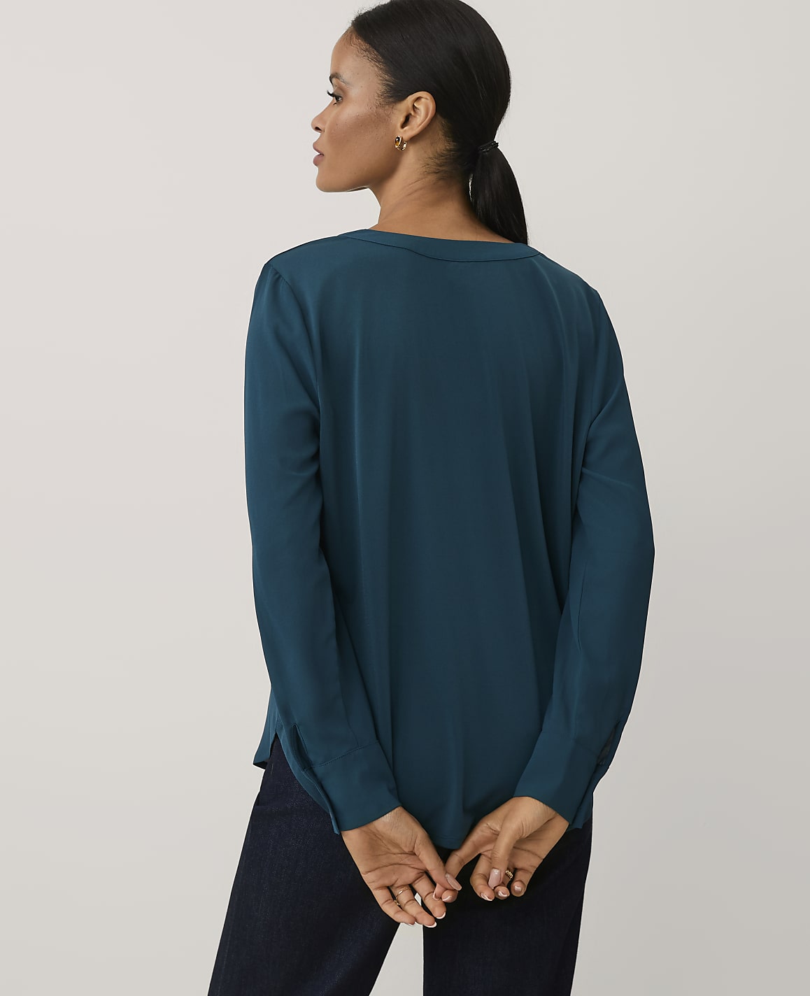 A woman wearing a teal long-sleeved blouse and dark trousers stands with her back to the camera against a plain light gray background.