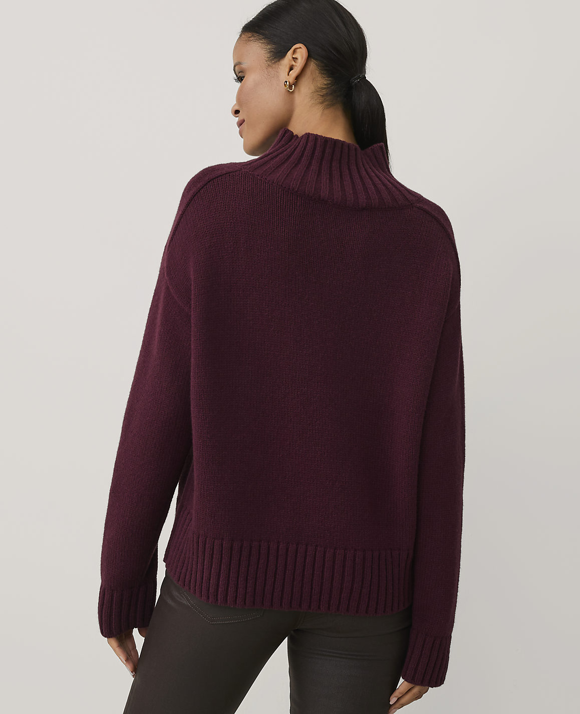A woman wearing a burgundy ribbed turtleneck sweater and dark brown pants stands against a plain light gray background.