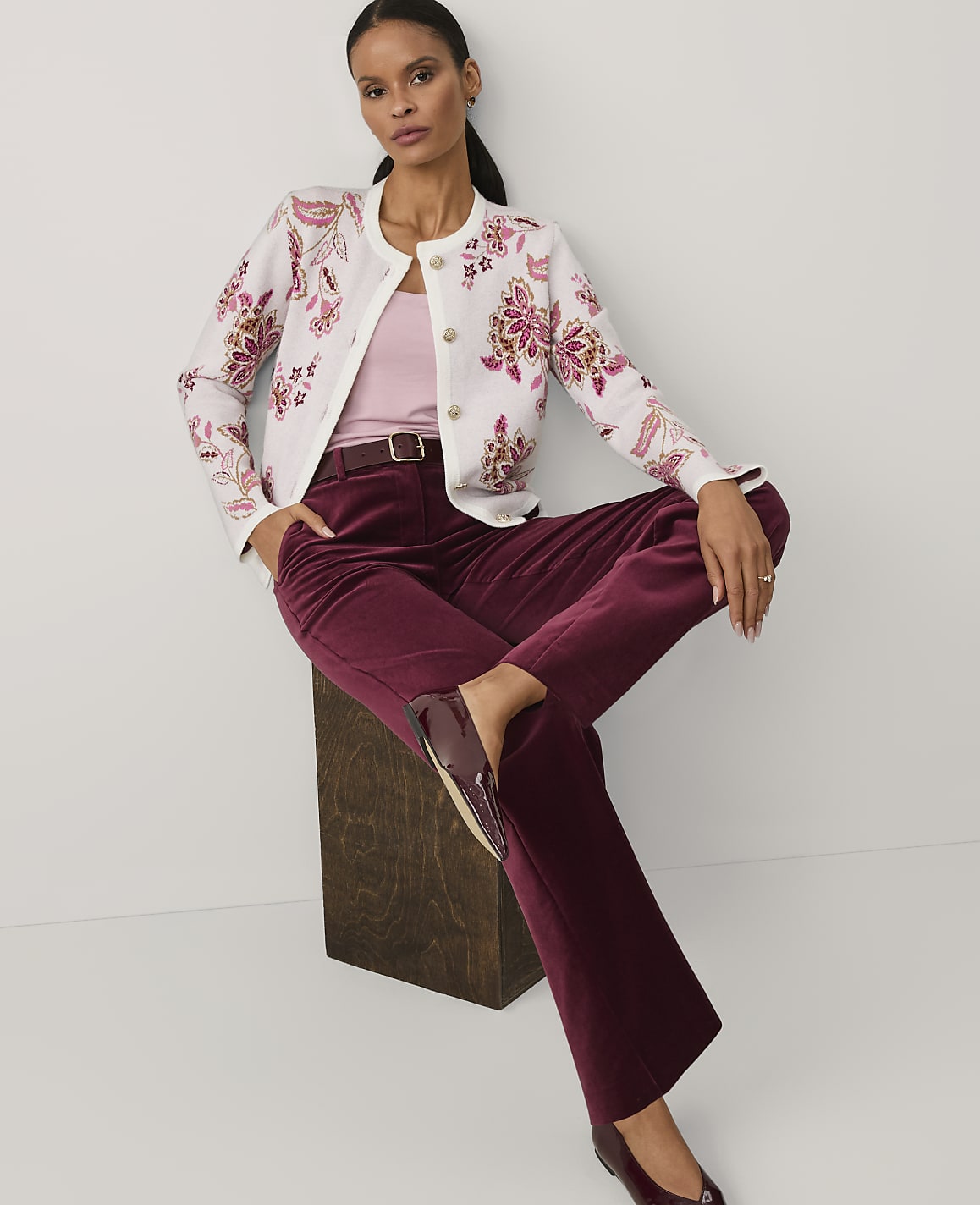 A woman wearing a floral white jacket and burgundy velvet trousers sits on a dark wooden block against a plain white background.