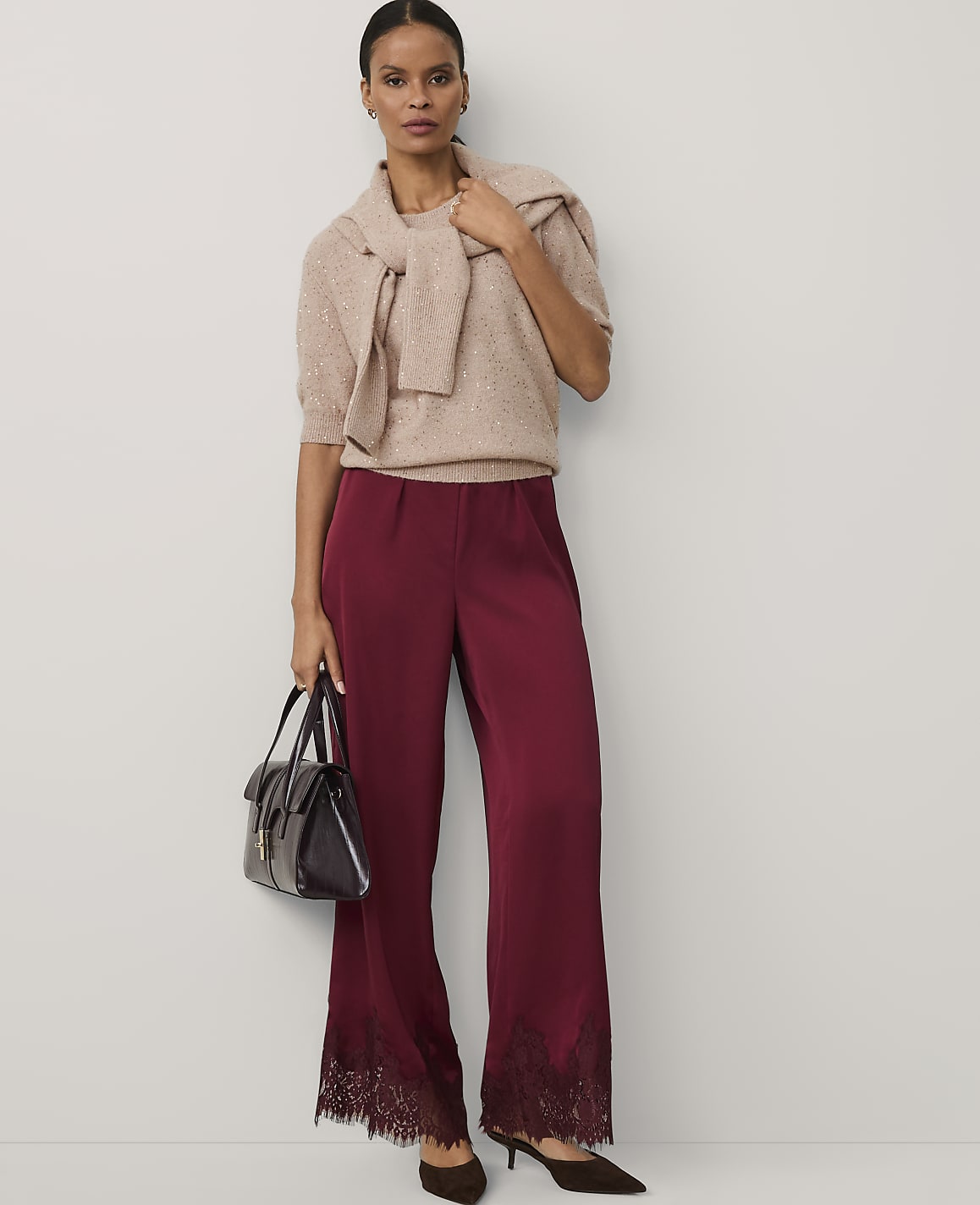 A woman wearing a beige sweater and burgundy lace-hemmed trousers stands against a plain white background.