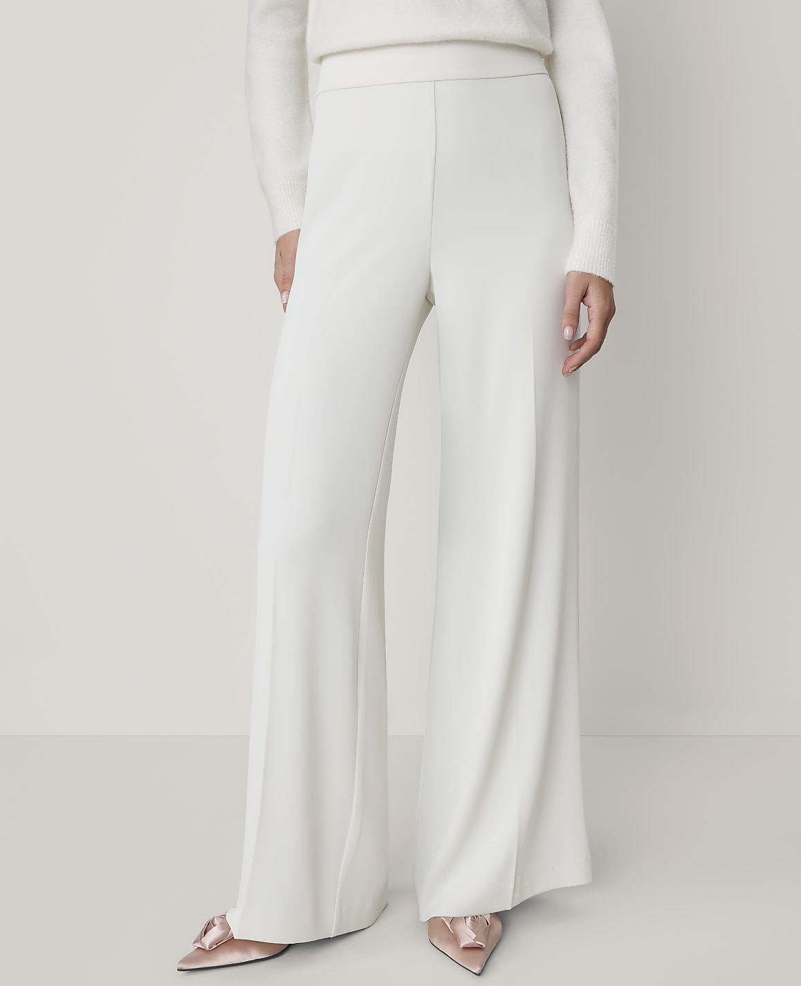 A person wearing a white sweater, wide-leg white trousers, and pointed pink heels stands against a plain, light-colored background.