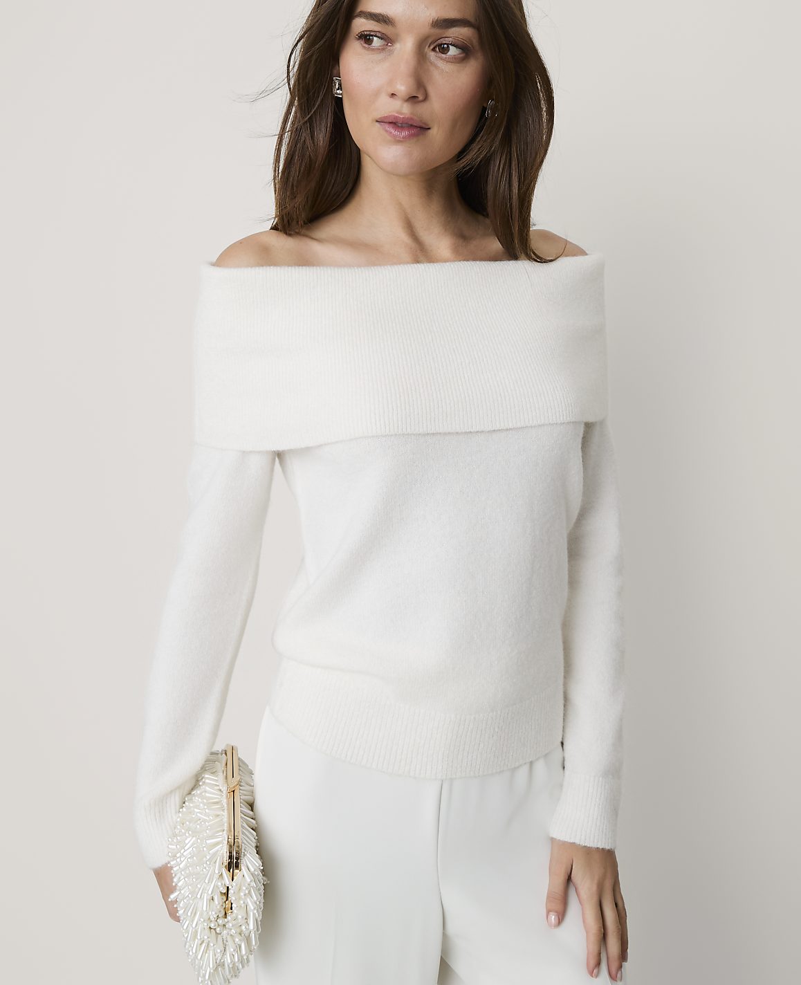 A woman wearing an off-the-shoulder white sweater and white trousers stands against a plain, light-colored background.