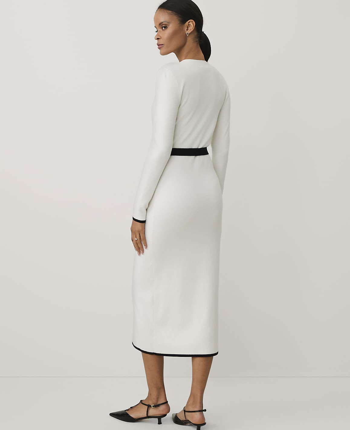 A woman wearing a long-sleeved white dress with black trim and black heels stands against a plain white background.