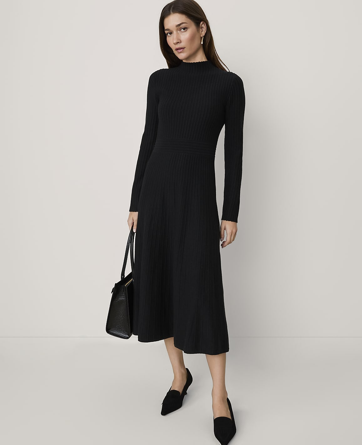 A woman wearing a long-sleeved black ribbed midi dress and black pointed-toe flats stands against a plain, light-colored background.