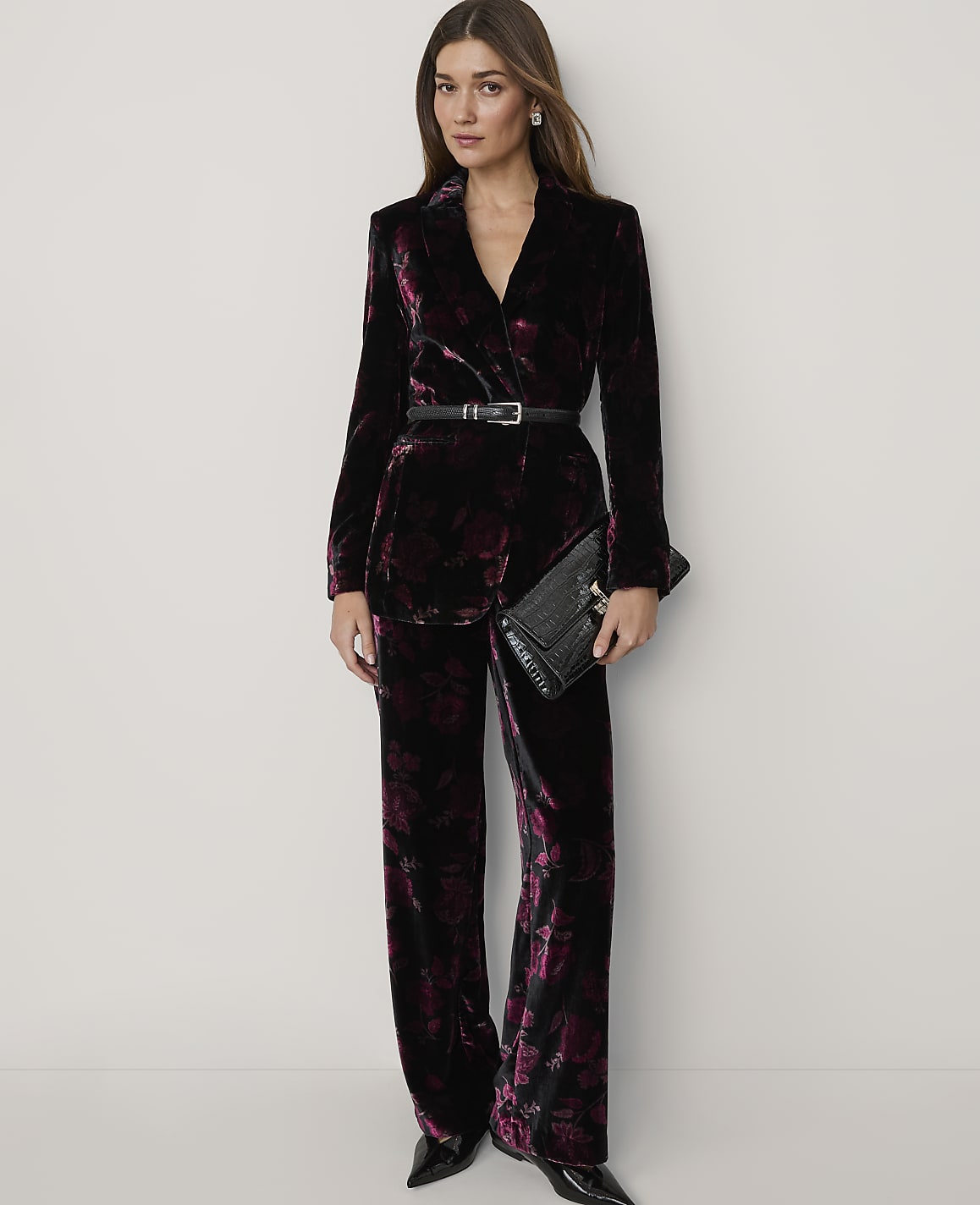 A woman wearing a dark purple velvet floral suit and holding a black clutch stands against a plain white background.