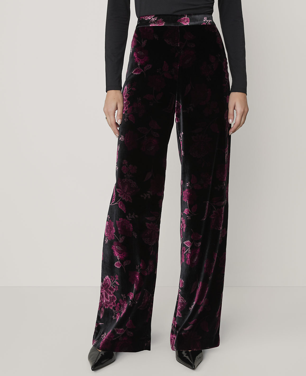 A person wears wide-leg black and purple floral velvet trousers and a black top against a plain light grey background.