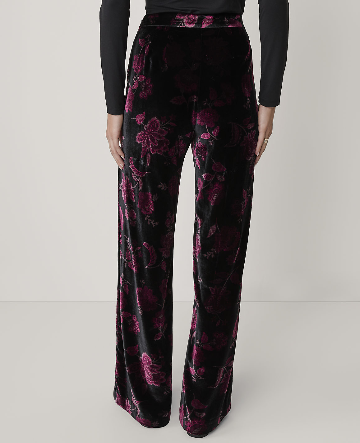 A person wears black long-sleeved top and wide-leg black velvet pants with a purple floral pattern against a plain light gray background.