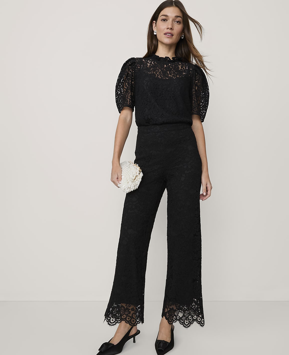A woman wearing a black lace jumpsuit and holding a white clutch stands against a plain, light-colored background.