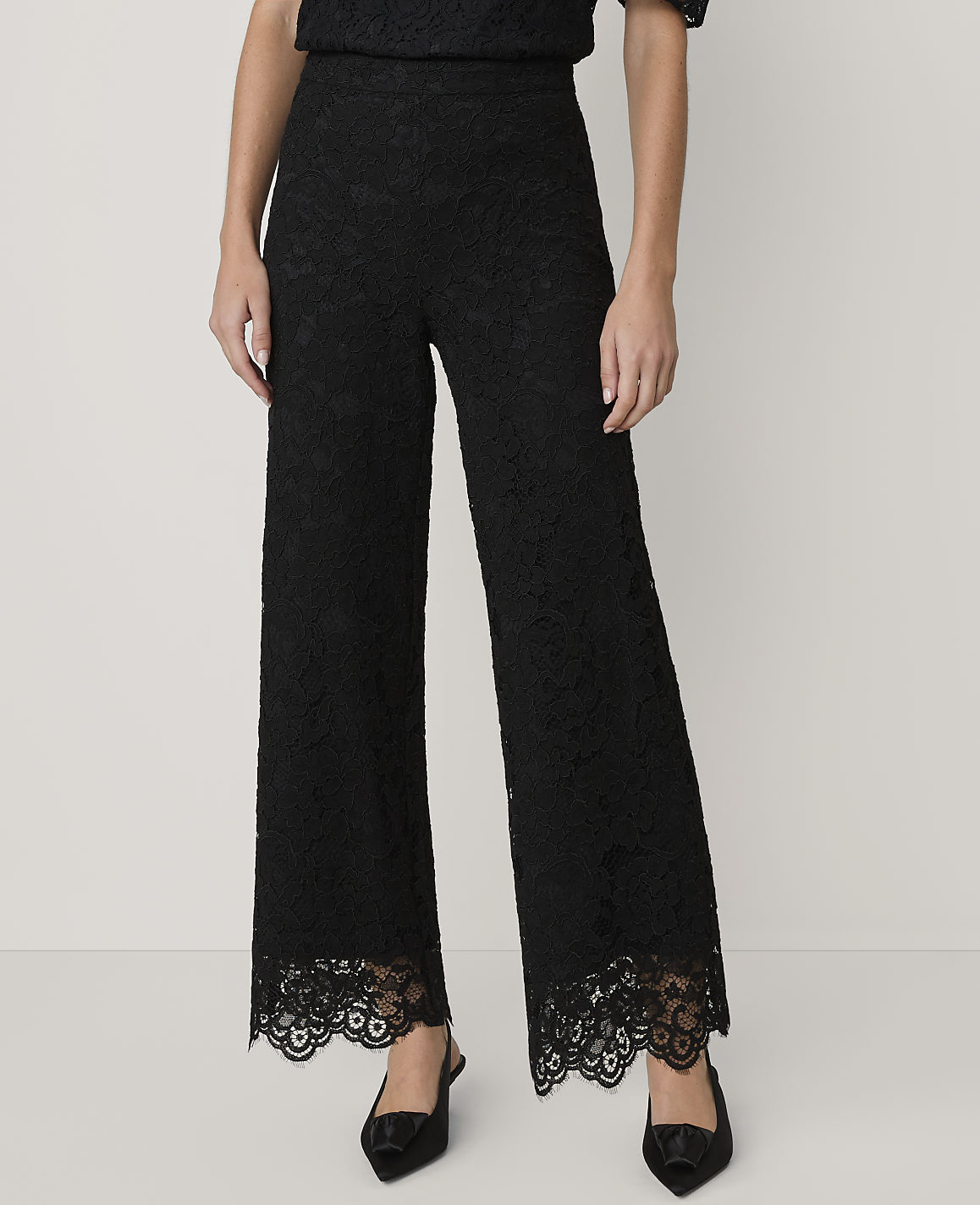 A person wears black lace wide-leg trousers and pointed heels against a plain light grey background.