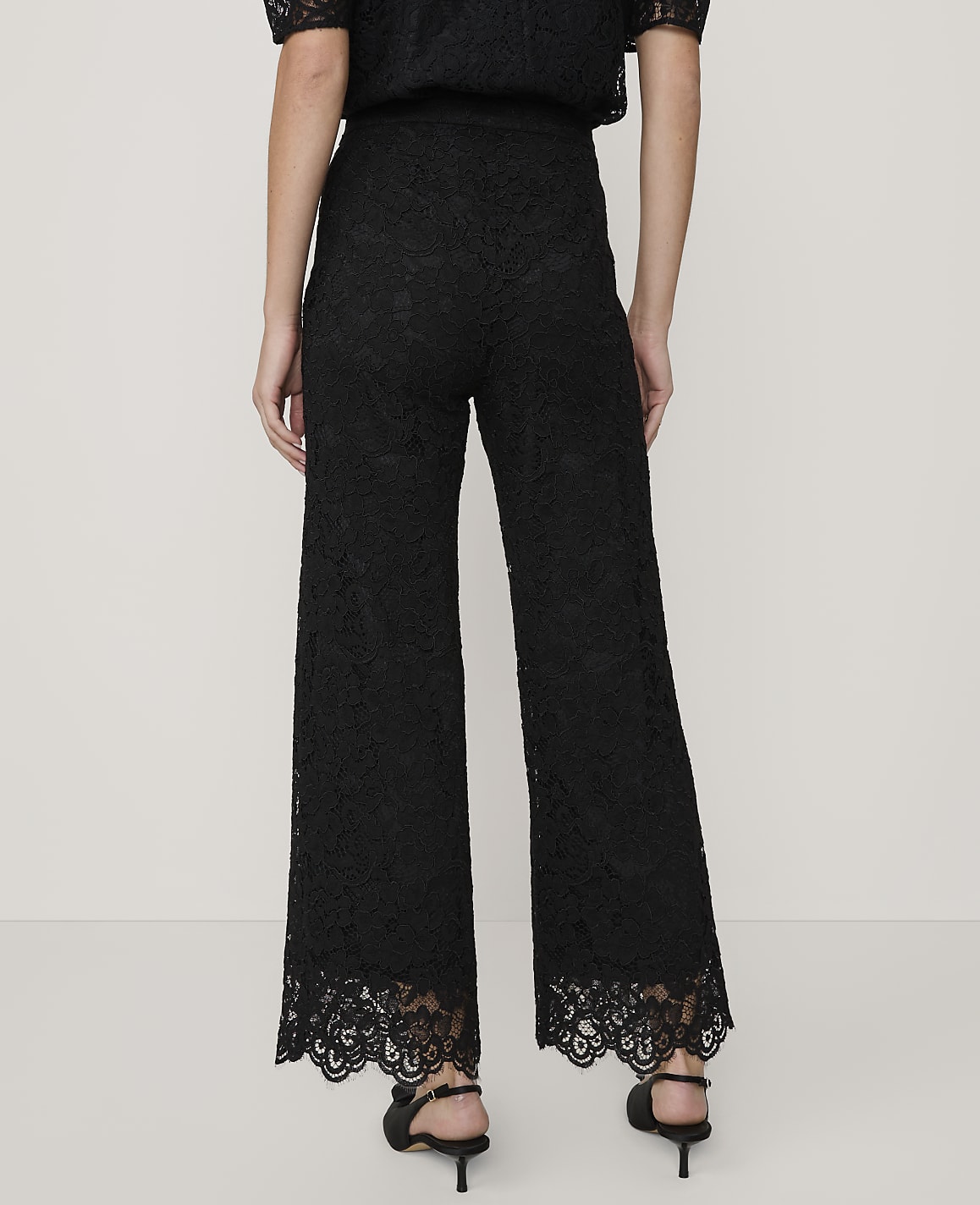 A person stands with their back to the camera wearing black lace wide-leg trousers and heels against a plain, light-colored background.