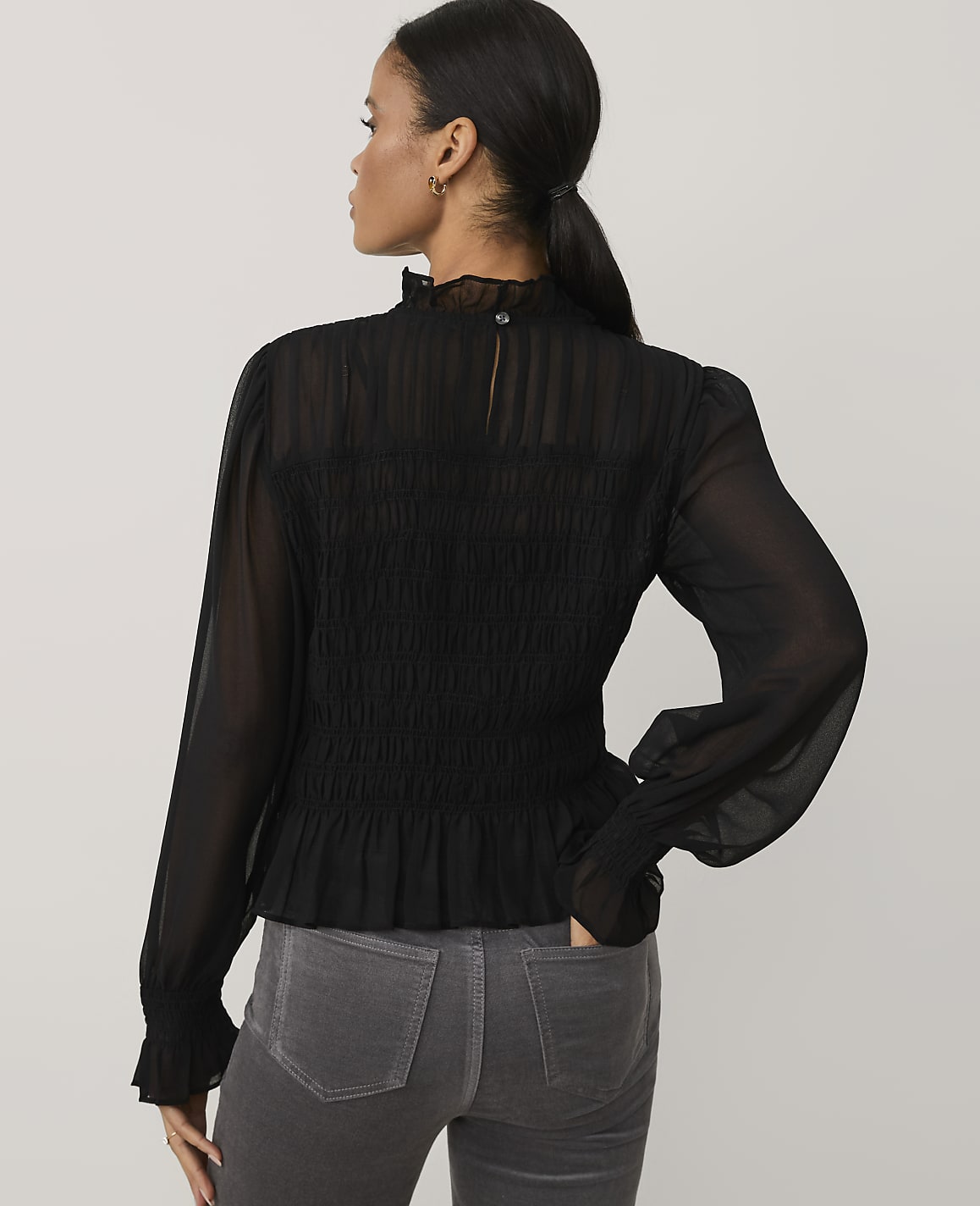 A woman wearing a black smocked blouse and grey jeans stands against a plain light grey background.