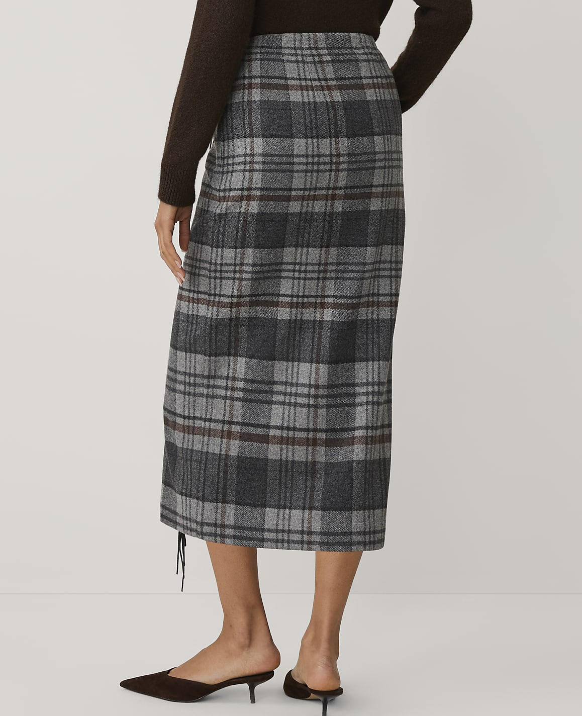 A person wearing a brown sweater, a grey plaid midi skirt, and brown pointed-toe heels stands against a plain light grey background.