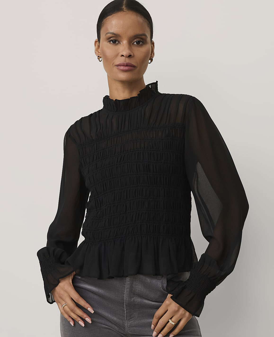 A woman wearing a black smocked blouse and grey trousers stands against a plain light grey background.