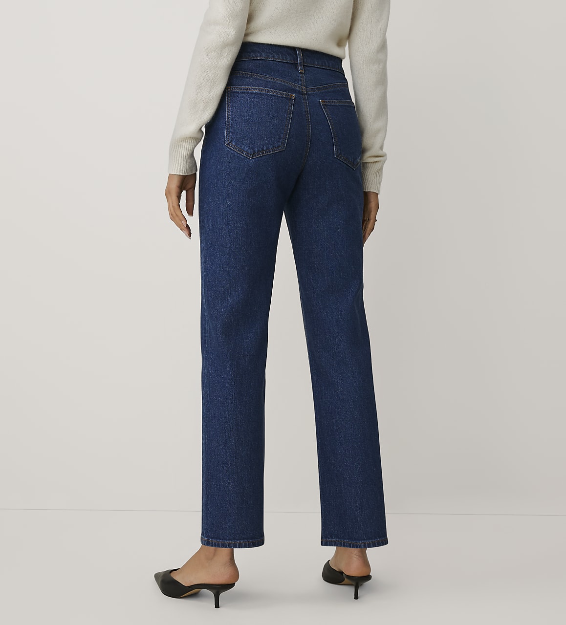 A person wearing a white sweater, blue jeans, and black mules stands against a plain white background.