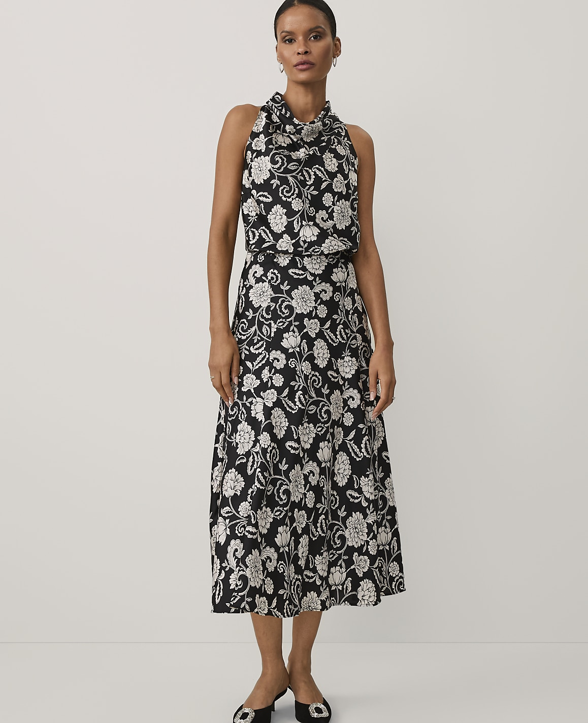 A woman wearing a black and white floral print dress and black heels stands against a plain white background.