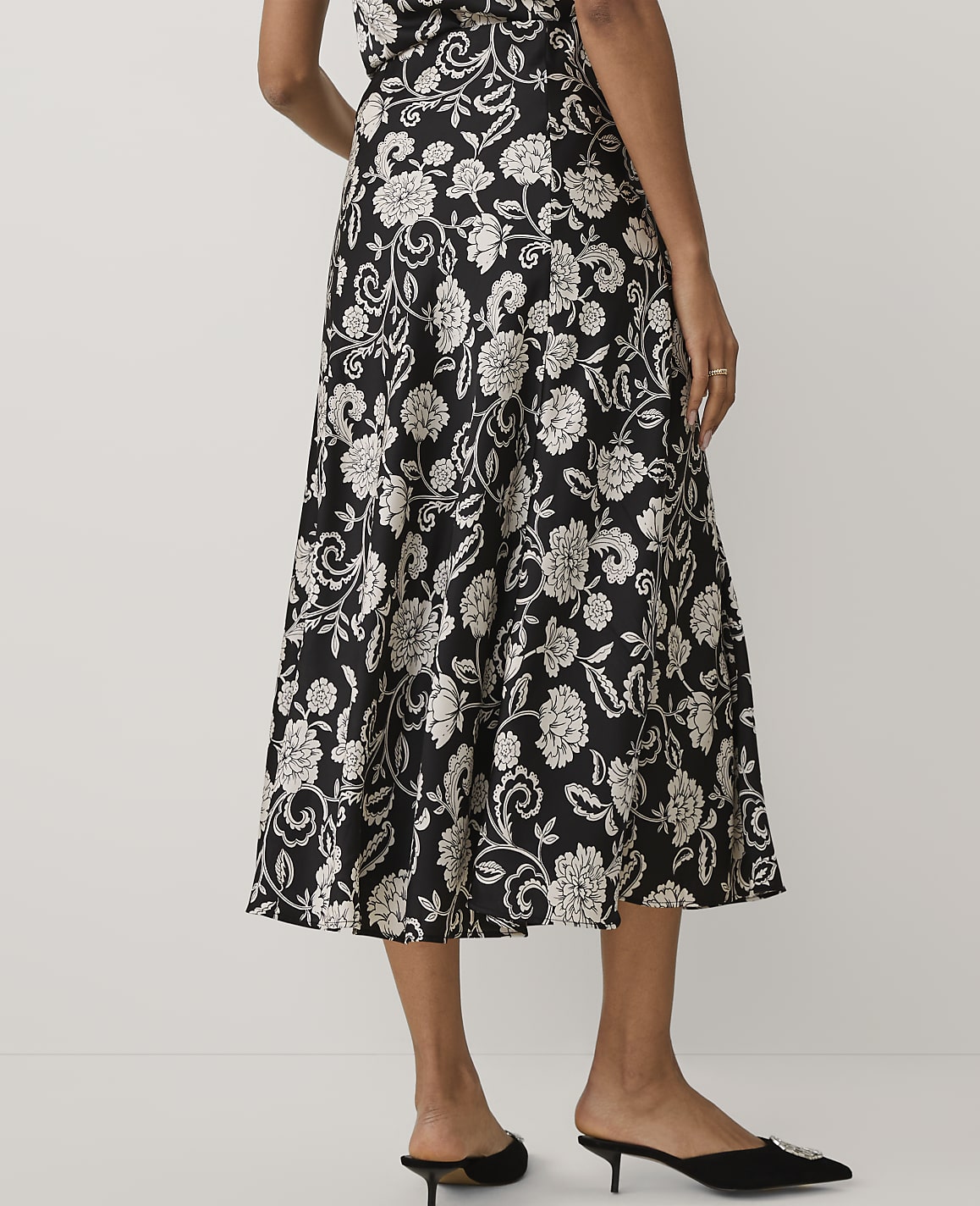 A person wears a black and white floral midi dress and black pointed-toe mules against a plain white background.