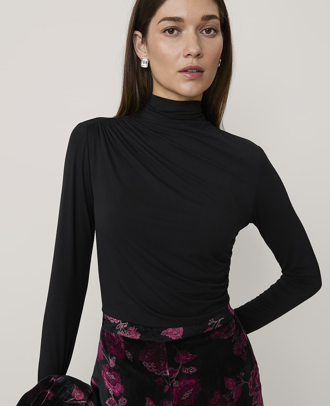 A woman wearing a black high-neck top and floral patterned trousers stands against a plain, light-colored background.