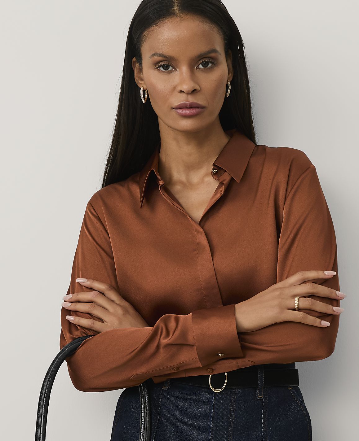 A woman with long dark hair wearing a copper-colored satin shirt and dark blue jeans stands with her arms crossed against a plain light gray background.