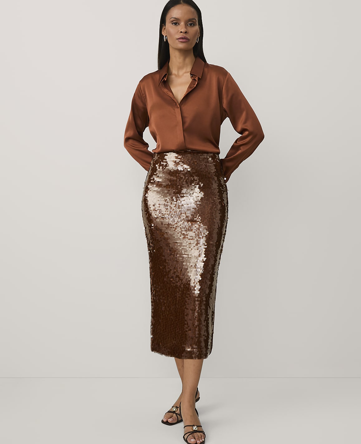 A woman wearing a brown satin shirt and a shimmering bronze sequined pencil skirt stands against a plain white background.