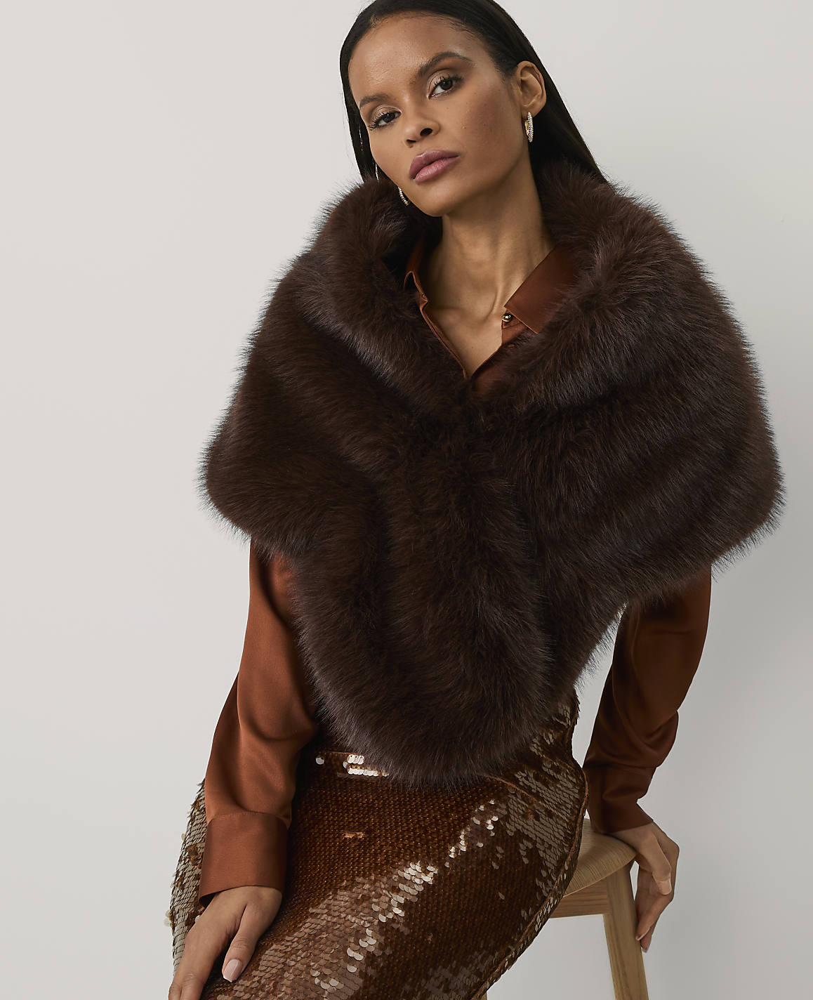 A woman wearing a brown satin shirt, a sequined skirt, and a plush brown fur stole poses against a plain white background.