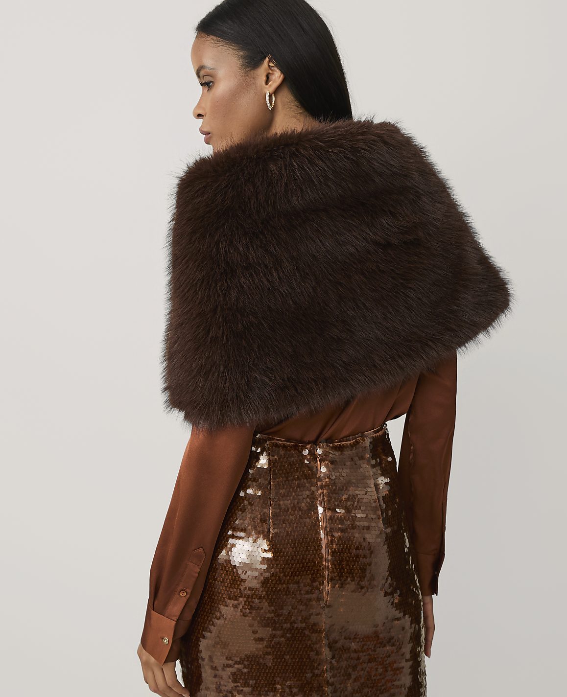 A woman wearing a brown faux-fur stole, a satin blouse, and a sequined skirt stands against a plain light gray background.