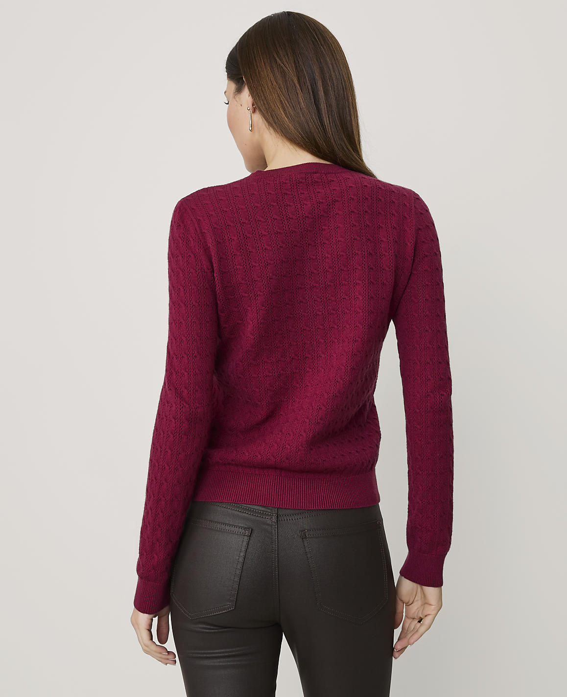 A woman wearing a textured magenta sweater and dark brown leather pants stands against a plain light gray background.