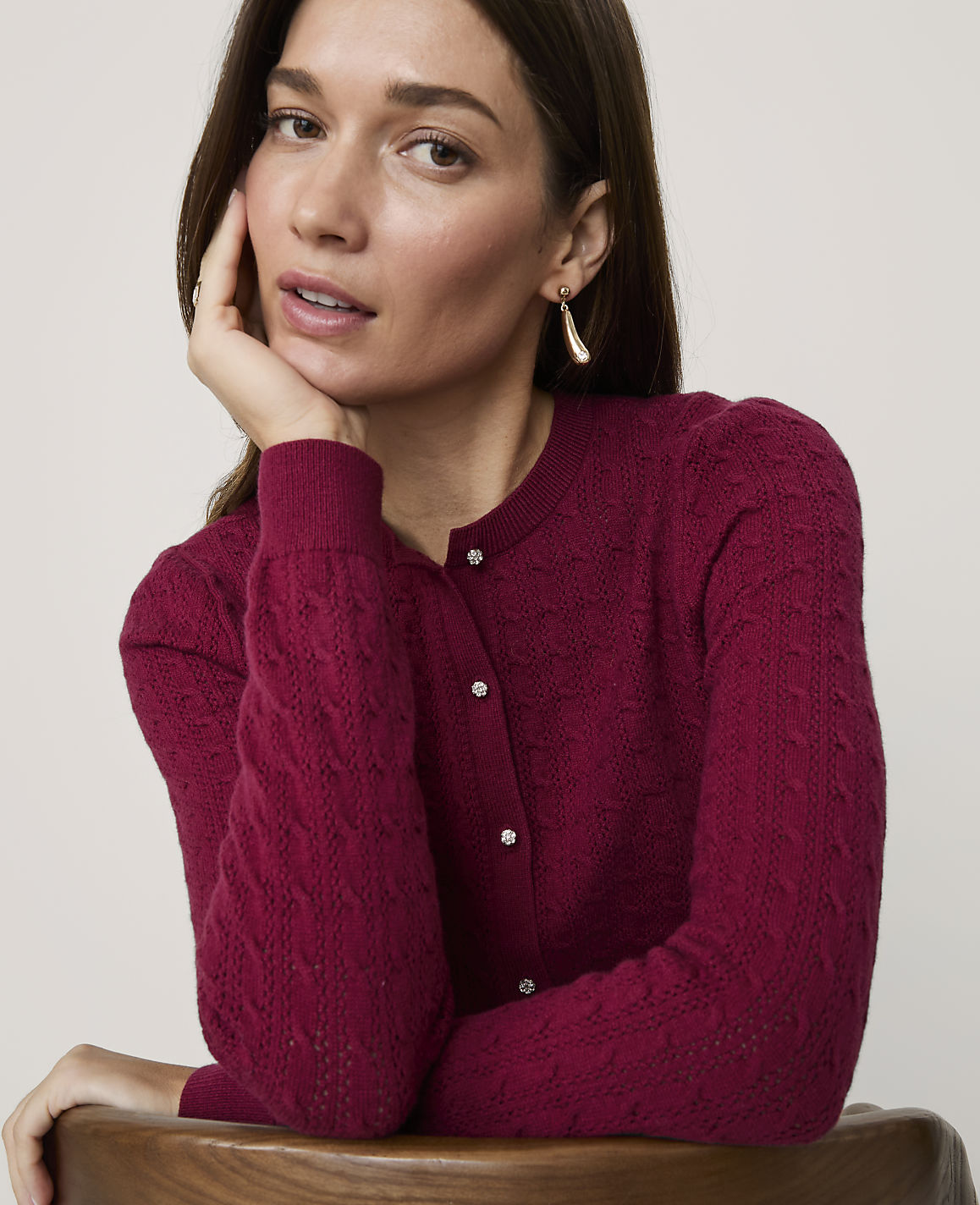 A woman in a textured magenta cardigan rests her chin on her hand against a plain, light-colored background.