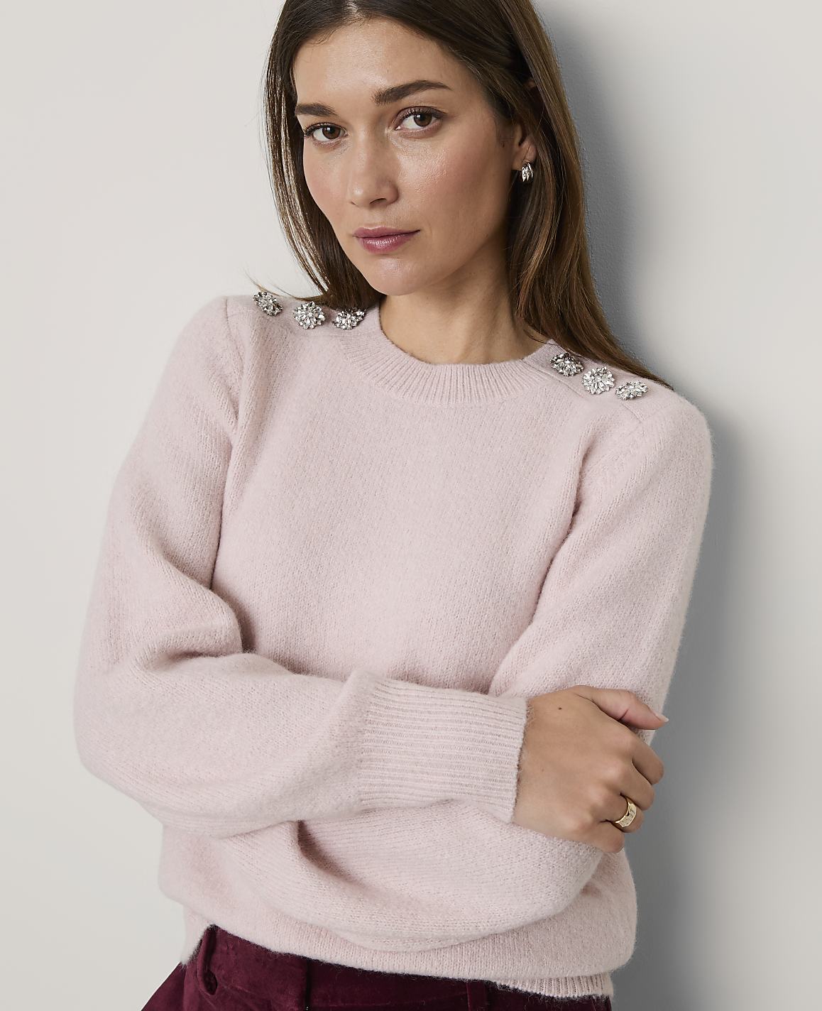 A woman wearing a light pink sweater with jeweled shoulder accents leans against a plain white wall.