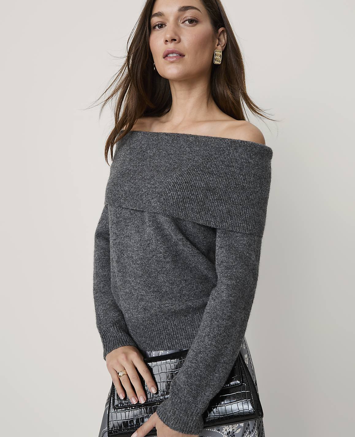 A woman wearing a grey off-the-shoulder sweater and holding a black clutch stands against a plain, light-colored background.