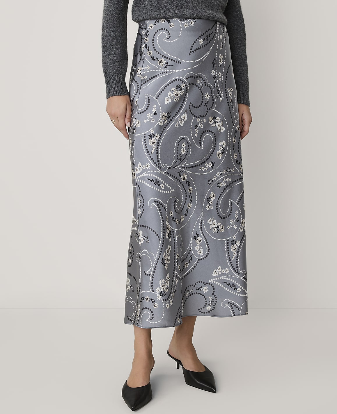A person wearing a grey sweater, a grey paisley-print midi skirt, and black pointed-toe heels stands against a plain white background.