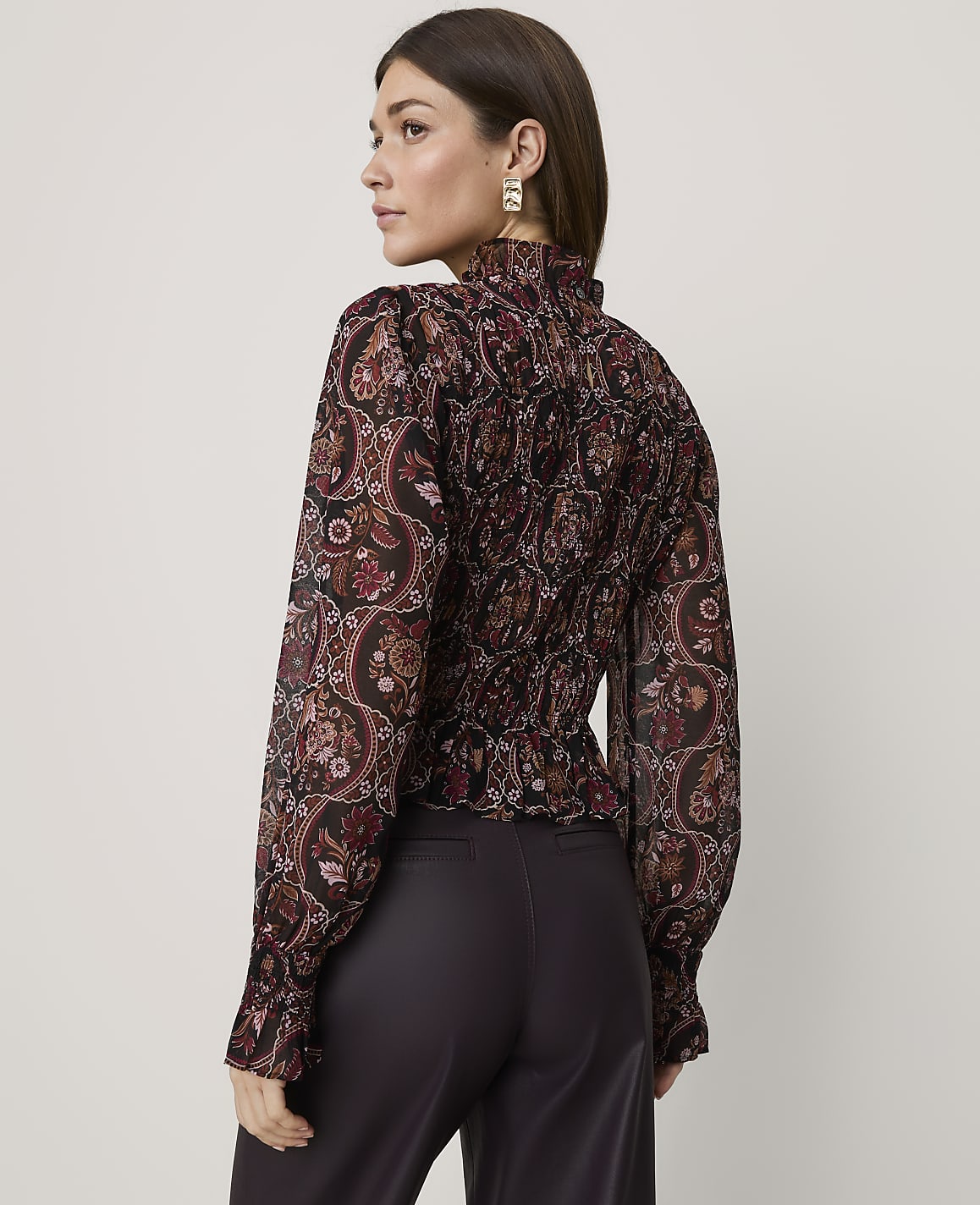 A woman wearing a patterned maroon blouse and dark trousers stands against a plain, light-colored background.
