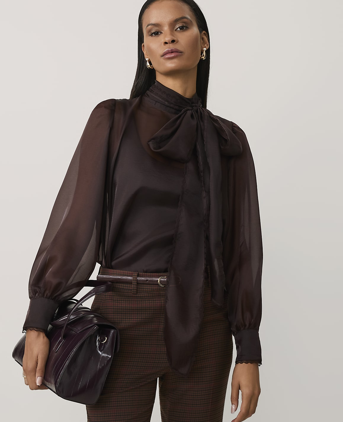 A woman wearing a sheer brown pussy-bow blouse and checkered trousers stands against a plain light grey background.