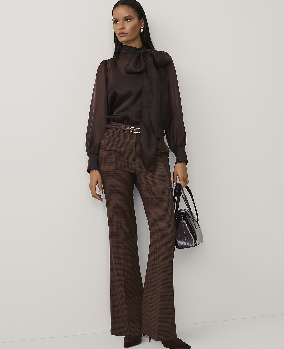 A woman wearing a brown pussy-bow blouse and plaid trousers stands against a plain white background.