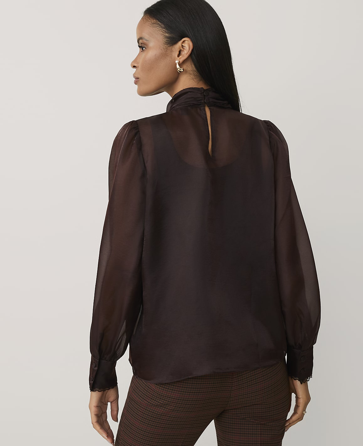 A woman wearing a sheer brown blouse and plaid trousers stands against a plain light gray background.