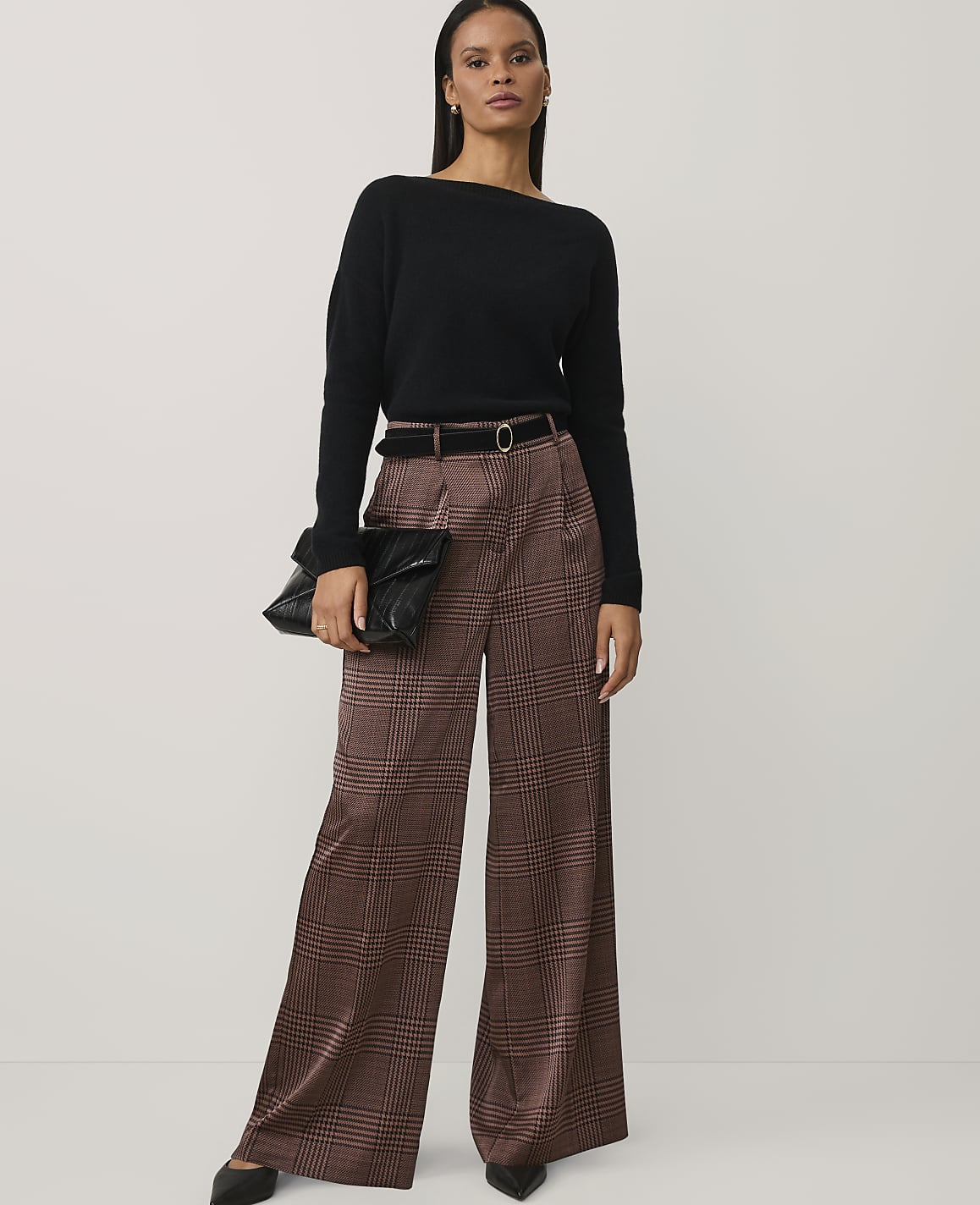 A woman wearing a black sweater and brown plaid wide-leg trousers stands against a plain, light-colored background.