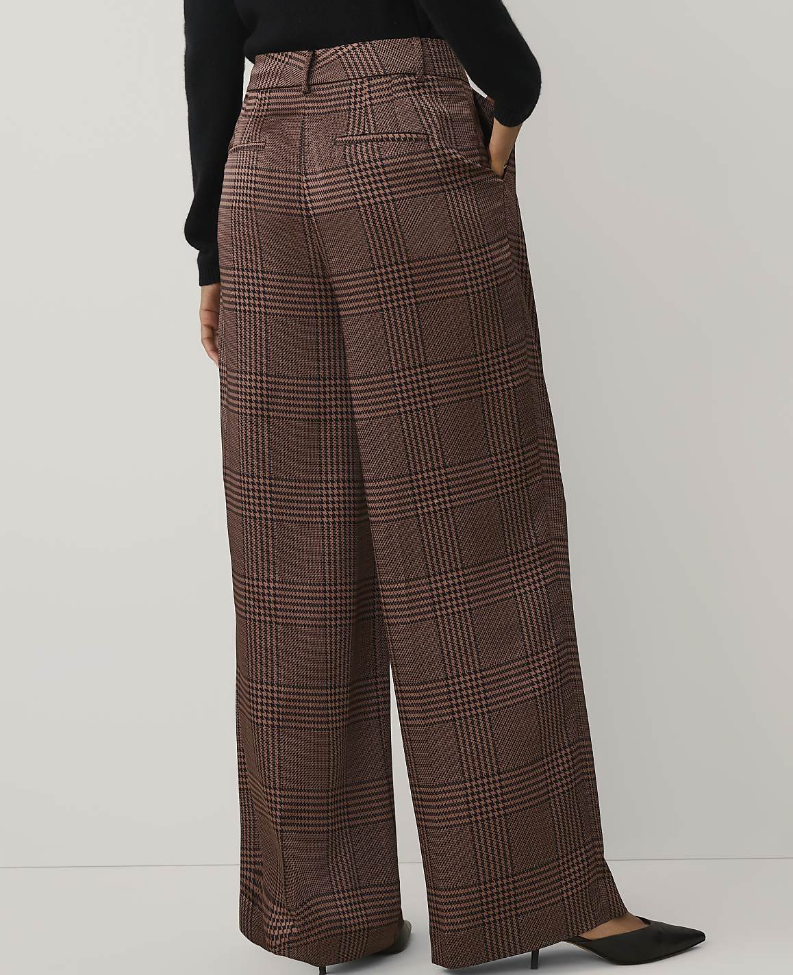 A person wearing a black top and brown plaid wide-leg trousers stands against a plain white background.
