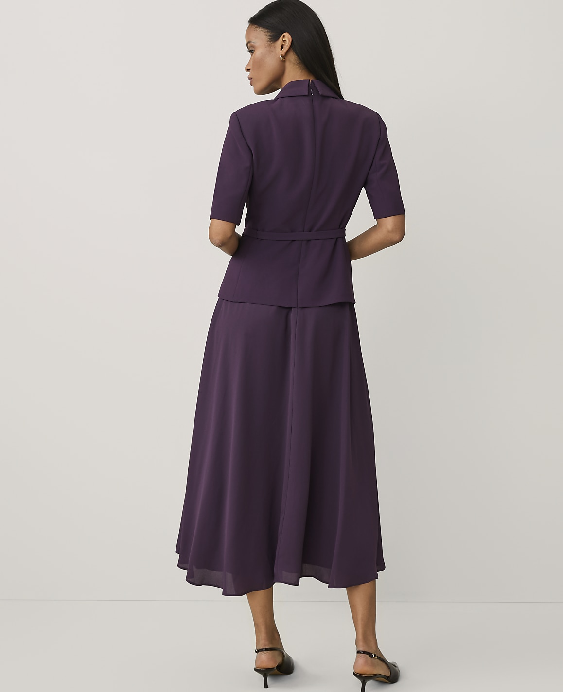 A woman stands with her back to the camera wearing a purple short-sleeved jacket and matching long skirt against a plain white background.