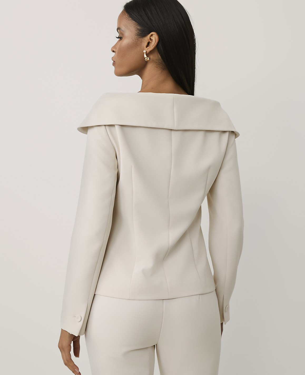 A woman wearing a cream-colored blazer and matching trousers stands with her back to the viewer against a plain, light-colored background.